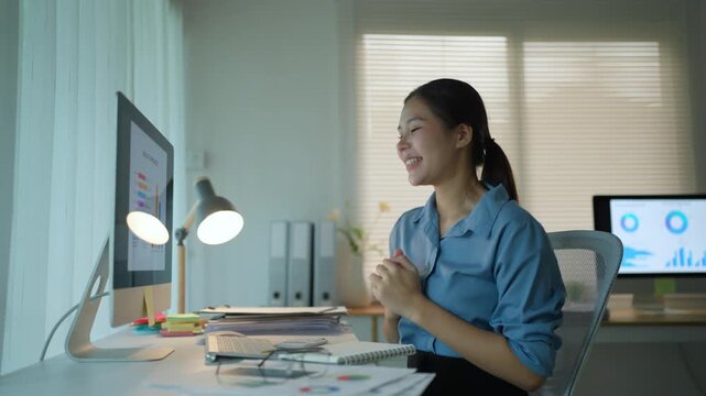 Joyful asian businesswoman celebrating a successful project completion, raising her fists and clapping with excitement while working late in the modern office at her computer desk