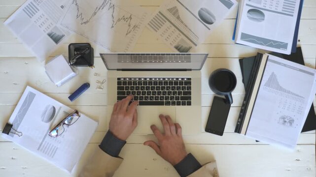 Male hands of manager typing text on laptop keyboard at white desk. Top view arms of young businessman looking at diagrams with statistics on papers and working on computer at wooden table in office