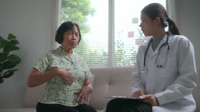 Senior woman suffering from abdominal pain discussing symptoms with a female doctor during a home visit, while the physician takes notes and provides a health consultation and check up