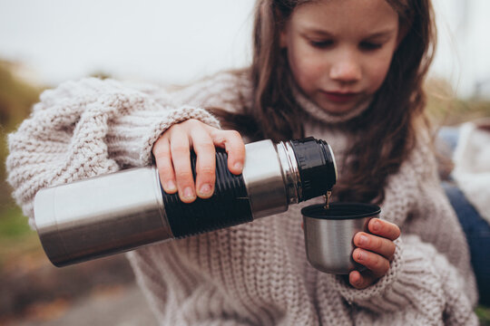 Young girl pouring hot drink from metal thermos into small cup outdoors. Autumn walk, family leisure time, outdoor adventure, simple joy of pausing for drink in nature.