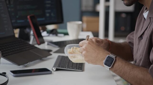 Handheld shot of focused young Indian man eating lunch from plastic container while working on laptop with code on screen. Ideal for software development, busy lifestyle, or IT industry marketing