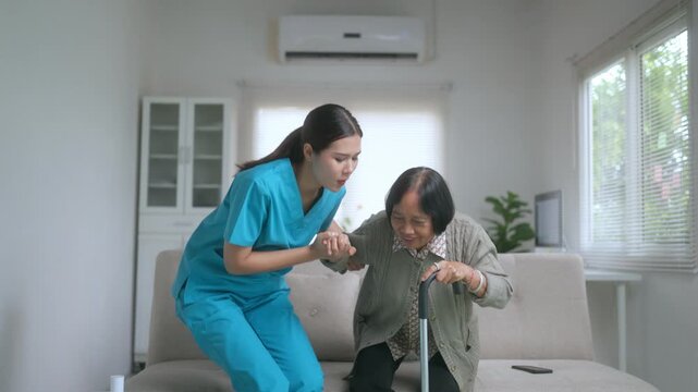 Compassionate young nurse in scrubs providing support to an elderly asian patient, helping her stand up from the sofa with a walking cane in the living room of a nursing home