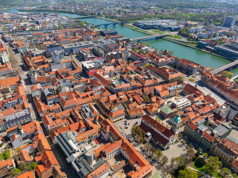 Aerial drone panorama displays historic downtown of Maribor city. Red terracotta roofs, old streets, and turquoise Drava river bridges shape urban landscape during sunny spring day.