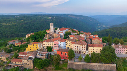 Fototapeta premium Drone captures medieval hilltop town of Oprtalj located in inland Istria. Historic stone houses, cypress trees, and ancient bell tower stand above picturesque countryside vineyards and fields.