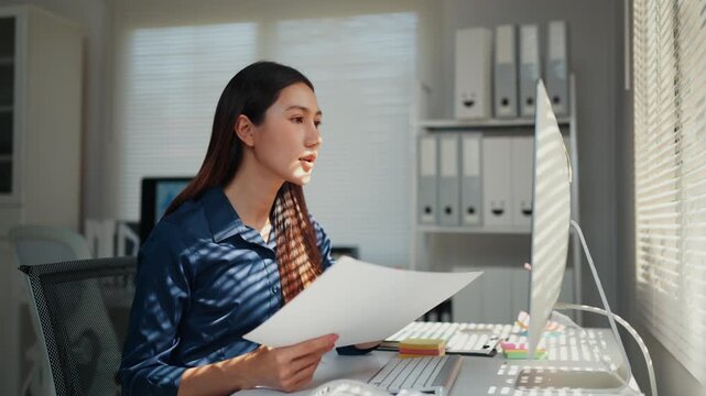 Professional asian businesswoman working at her desk, holding a paper document while pointing at the computer screen with a pen to compare information in a sunlit modern office