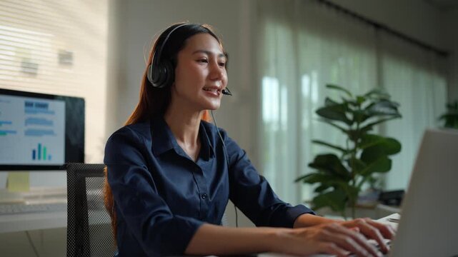 Young Asian woman wearing a headset working as a customer service representative, talking and smiling while looking at a laptop computer screen in her home office during the evening