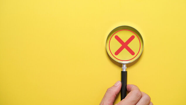 Hand holding a magnifying glass highlighting a red cross symbol for concept of error detection, rejection of quality control, problem identification, risk assessment, compliance audit and inspection.