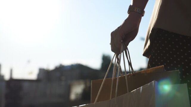 Female arm holds full paper packets walking along urban street. Hand of young woman carries shopping bags going on city alley after purchases. Concept of seasonal sales and discounts. Close up Slow mo