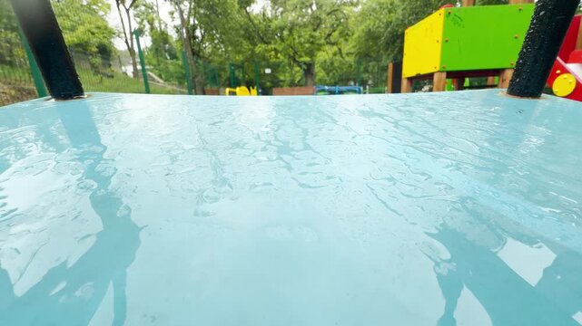a wet blue swing seat moves slowly in the wind. The motion creates a soft metallic squeaking sound. Water droplets drip quietly from the edge.