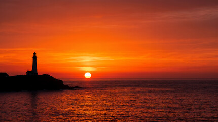 Fototapeta premium Silhouette of a coastal lighthouse on a rocky cliff during a vibrant fiery sunset over the ocean