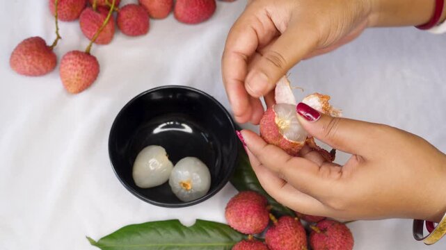 A woman hand Holding a Freshly Peeled Lychee Fruit, Revealing Its Juicy White Flesh and red Outer Shell on keep black bowl. inside with another whole lychee or litchi fruits.