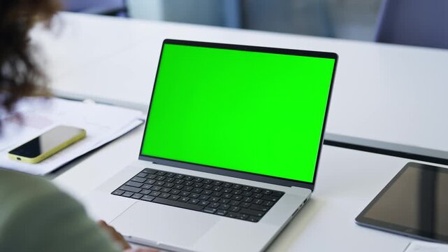 Over shoulder view of female person hand using laptop with green screen chroma key mockup at desk table in modern office for business, work, education. Space for advertising, advertisement, ecommerce
