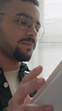 Vertical shot of Hispanic young adult male student wearing glasses, opening book and reading pages attentively. Ideal for educational marketing, library promotions, and diversity campaigns