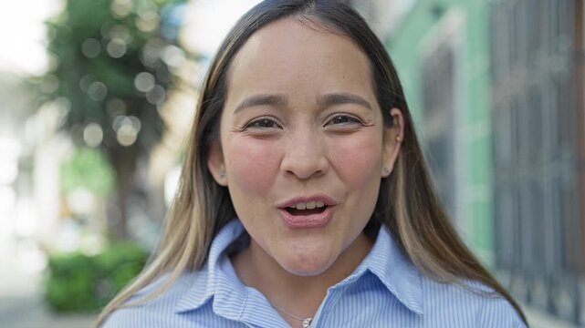 Young hispanic woman mouth open smiling with visible teeth and rosy cheeks on a city street closeup, wearing blue striped shirt and necklace; joy warmth.