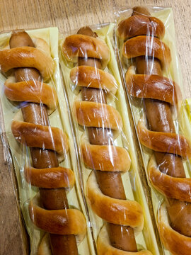 Three long hot dog sausages neatly wrapped in a spiral layer of golden-brown baked dough, each individually packaged in long, clear plastic sleeves laid out on a wooden surface.