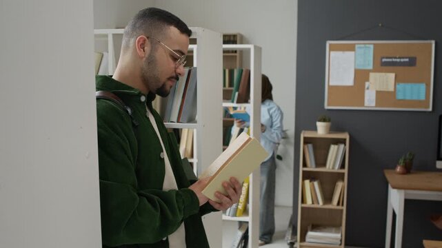 Middle Eastern young adult male student reading book in library while female classmate in hijab browses shelves in background. Video is ideal for university diversity campaigns