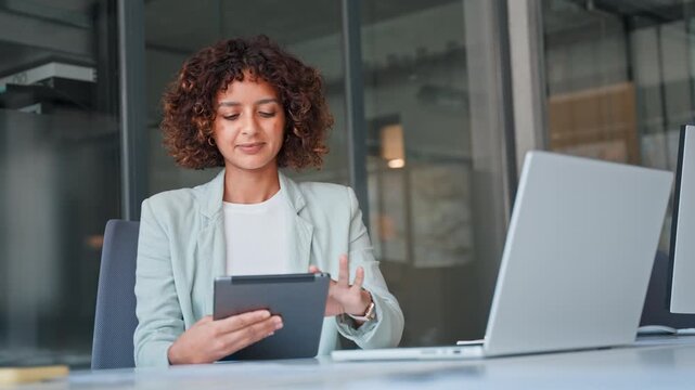 Arabian young businesswoman in blazer sitting at office desk, using tablet and laptop in modern workspace. Professional indian woman bank worker working with technology fintech device for business
