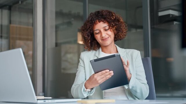 Smiling latin businesswoman sitting at office desk and using tablet at laptop in modern workspace, communication. Young indian woman holding tab screen device for work online. Technology and business