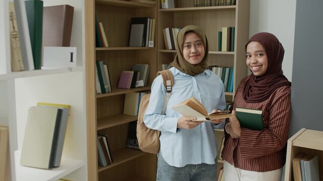 Portrait of two Asian Muslim young adult female students holding books and smiling in library. Video is perfect for marketing campaigns on diverse education and international student programs