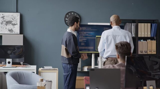 Medium long shot of young Indian male programmer discussing code with Middle Eastern colleague presenting software during office meeting. For IT corporate training and software development marketing