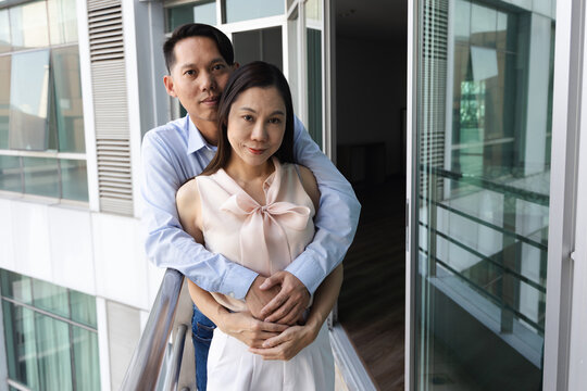 Asian husband embraces wife from behind at balcony, concept of happy couple family life