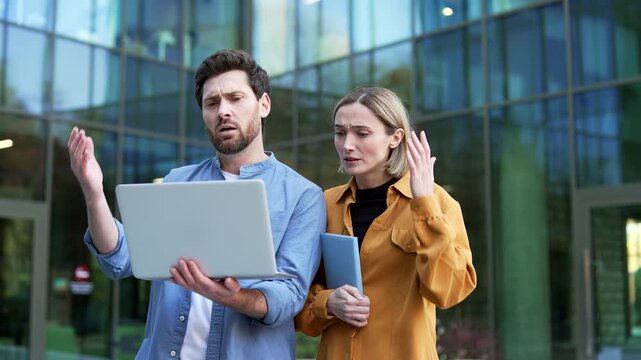 Businessman and businesswoman review unfavorable information on a laptop standing near office building. Concerned colleagues react to business difficulties, financial losses and disappointing results