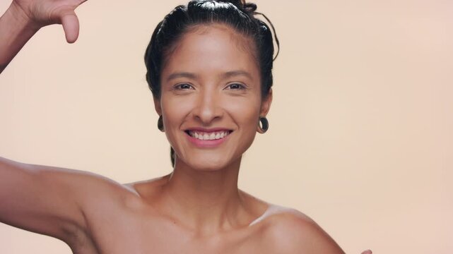 Close-up shot of argentinian woman with honey skin tone makes finger frame, smiling illustrating pure skin over tan background | Beauty ad concept