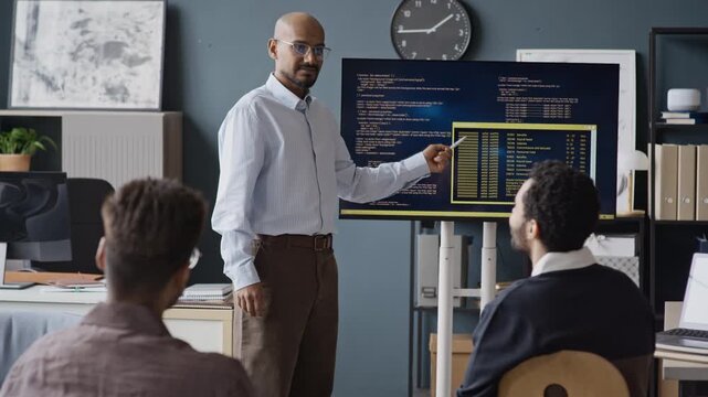 Slowmo shot of young Indian male IT specialist pointing at code on screen, presenting software development to diverse colleagues. Ideal for corporate training and IT business presentations
