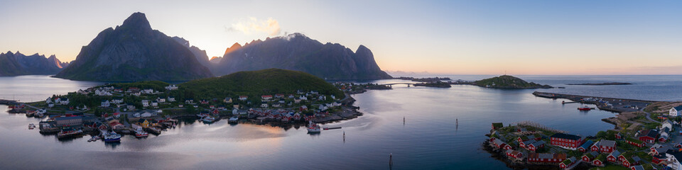 Aerial panorama shows Reine and Reinebringen in Lofoten, Norway. Red and white cabins sit on islets linked by bridges, buoys dot calm fjords at pastel twilight. © Aerial Film Studio