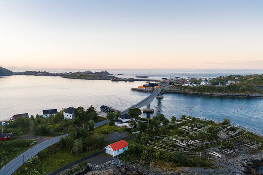 Aerial view shows Lofoten, Norway, a coastal village with a bridge over turquoise water, red and white houses on green slopes, hjell racks, quiet roads, and long evening shadows.
