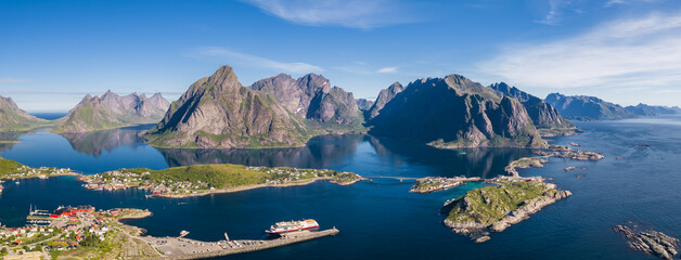 Aerial view of Reine and Reinebringen in Lofoten, Norway. Jagged peaks rise from fjords, a cruise ship docks, red and white cabins line islets with low bridges. © Aerial Film Studio