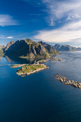 Fototapeta premium Aerial view of Lofoten Islands in northern Norway shows granite peaks above deep fjords, rocky islets, Reine and Reinebringen area, a coastal settlement, cabins, road, summer daylight