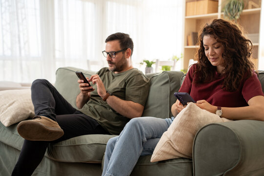 Angry young couple ignoring each other using phone after an argument while sitting on sofa at home.
