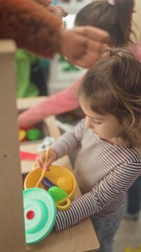 Girl stirs colorful plastic toy pot with a spoon, hand visible, another child watches in a daycare building play kitchen; playful curiosity.