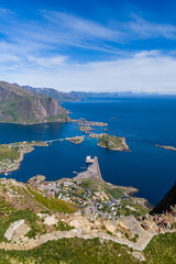 Obraz premium Aerial view shows Reine, harbor causeway, red and white cabins, marina, ferry, winding roads, steep rocky mountains, emerald slopes, islets, and summer Arctic light.