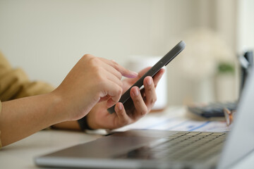 Fototapeta premium Close up of hands using smartphone beside laptop, calculator, and financial documents in a home office.