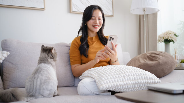 Smiling young woman sitting on a sofa at home with a cat beside her, using a smartphone in a cozy living room.