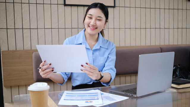 Young businesswoman writing notes on financial documents while working on a laptop in a modern cafe.