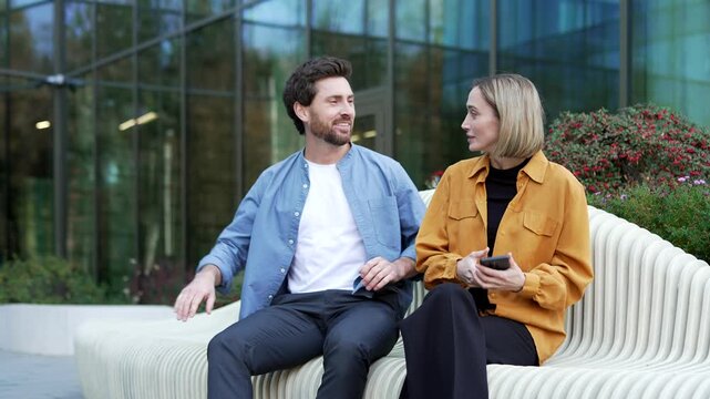 Businessman makes unwanted advances toward a businesswoman sitting on a bench near office building. Female colleague appears annoyed, uncomfortable as inappropriate workplace behavior unfolds outdoors