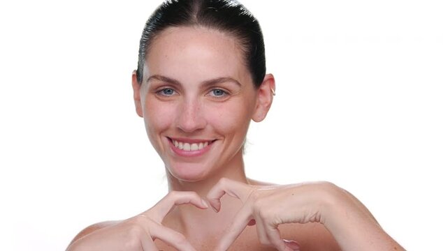 Close-up of young fair-skinned Latin woman with blue eyes and ponytale hairstyle showing hand heart gesture on white bg | Skin care concept