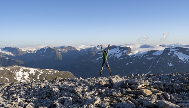 Mountain panorama with mountain peaks and glacier Jostedalsbreen, mountaineer stretches his arms in the air and jumps, at the summit of Sk&aring;la, Loen, Norway