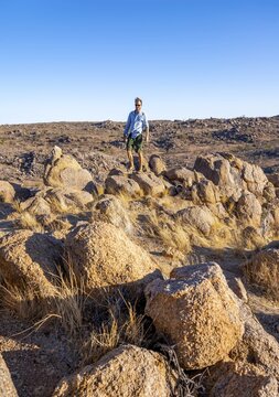Young man on a rockScarce landscape with rocky hills and acacia trees, African savannah in the evening light, Hobatere Concession, Kunene, Namibia