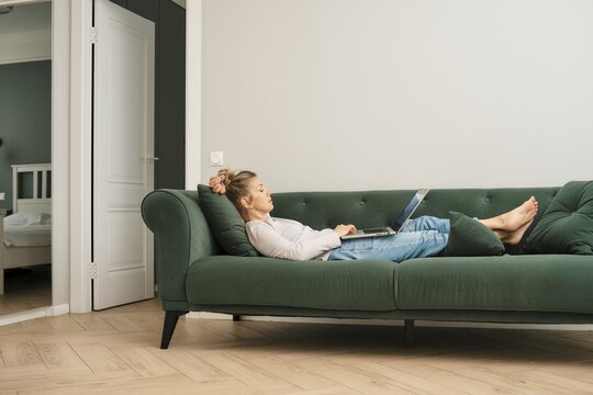 Middle-aged woman is leaning back comfortably on a green sofa, deeply focused on her laptop. With a casual outfit and an inviting living space, she is enjoying her time alone at home