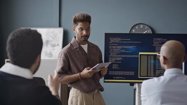 Handheld shot of confident young Indian male IT specialist presenting code on screen to multiethnic colleagues in office. Clip is suitable for software development and corporate training videos