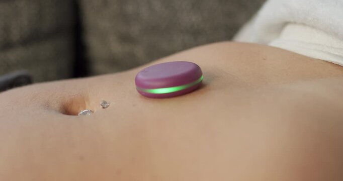 Close up of a round purple device with a green glowing ring resting on the belly of a person