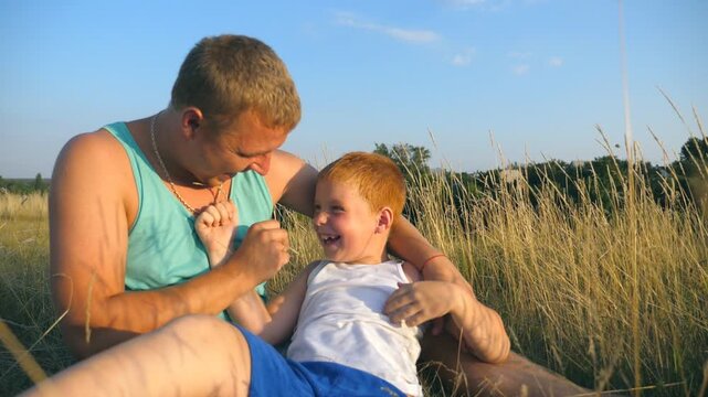 Father tickling his little son outdoor. Happy young man lying on grass at the field and playing with his baby boy. Cheerful daddy and his child having fun together in a park. Slow motion Close up