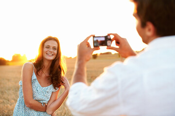 Obraz premium Woman, sunset and camera with couple in meadow, photography or memory of honeymoon. Female person, smile and nature with man and digital tech for picture taking, anniversary trip and relationship