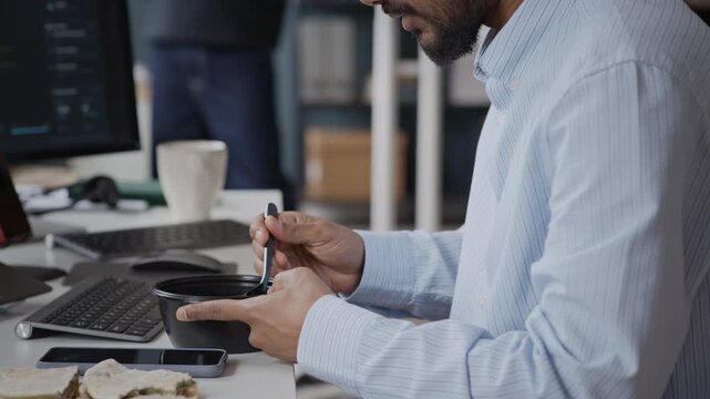 Tilt up shot of Indian male programmer eating lunch at office desk while looking at computer screens with code. Perfect for videos on software development, busy corporate life, or multitasking