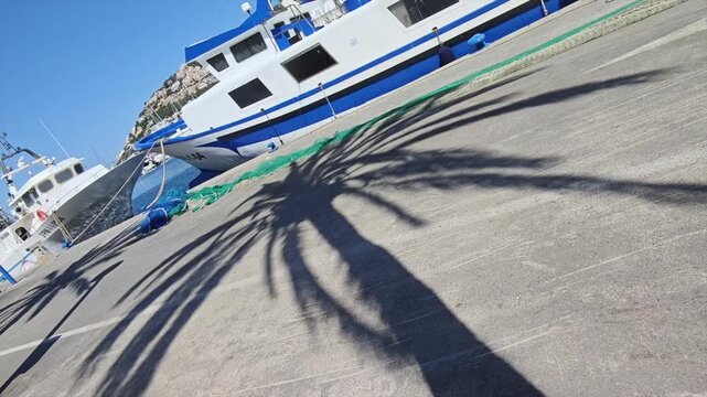 Palm Tree Shadow Over Fishing Nets on Coastal Street with Boat and Sea in Background