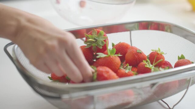 Strawberries are poured from glass bowl into metal sieve. Concept of washing fruit, fresh berries and summer food preparation